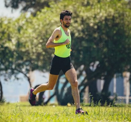 Full length of young bearded sportsman in activewear running on grassy lawn in city park near green trees during outdoor workout