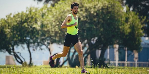 Full length of young bearded sportsman in activewear running on grassy lawn in city park near green trees during outdoor workout